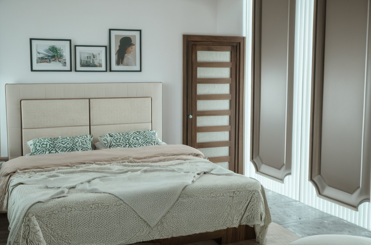 Jasna sypialnia z beżowym tapicerowanym zagłówkiem, wzorzystymi poduszkami i narzutą o wyraźnej fakturze, uzupełniona drewnianymi drzwiami z przeszkleniami i dekoracyjnymi panelami ściennymi.