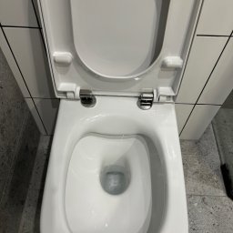 Firma sprzątająca Klaudia Dudek - Widok z góry na otwartą, białą, wiszącą toaletę ze spłuczką, zamontowaną na tle białych płytek z czarnymi fugami i szarej posadzki.