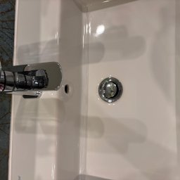 Firma sprzątająca Klaudia Dudek - Biała umywalka z chromowaną baterią i korkiem spustowym na tle ciemnej, wzorzystej ściany.