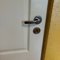 Białe drzwi z chromowaną klamką i zamkiem na tle ż&oacute;łtej, teksturowanej ściany.