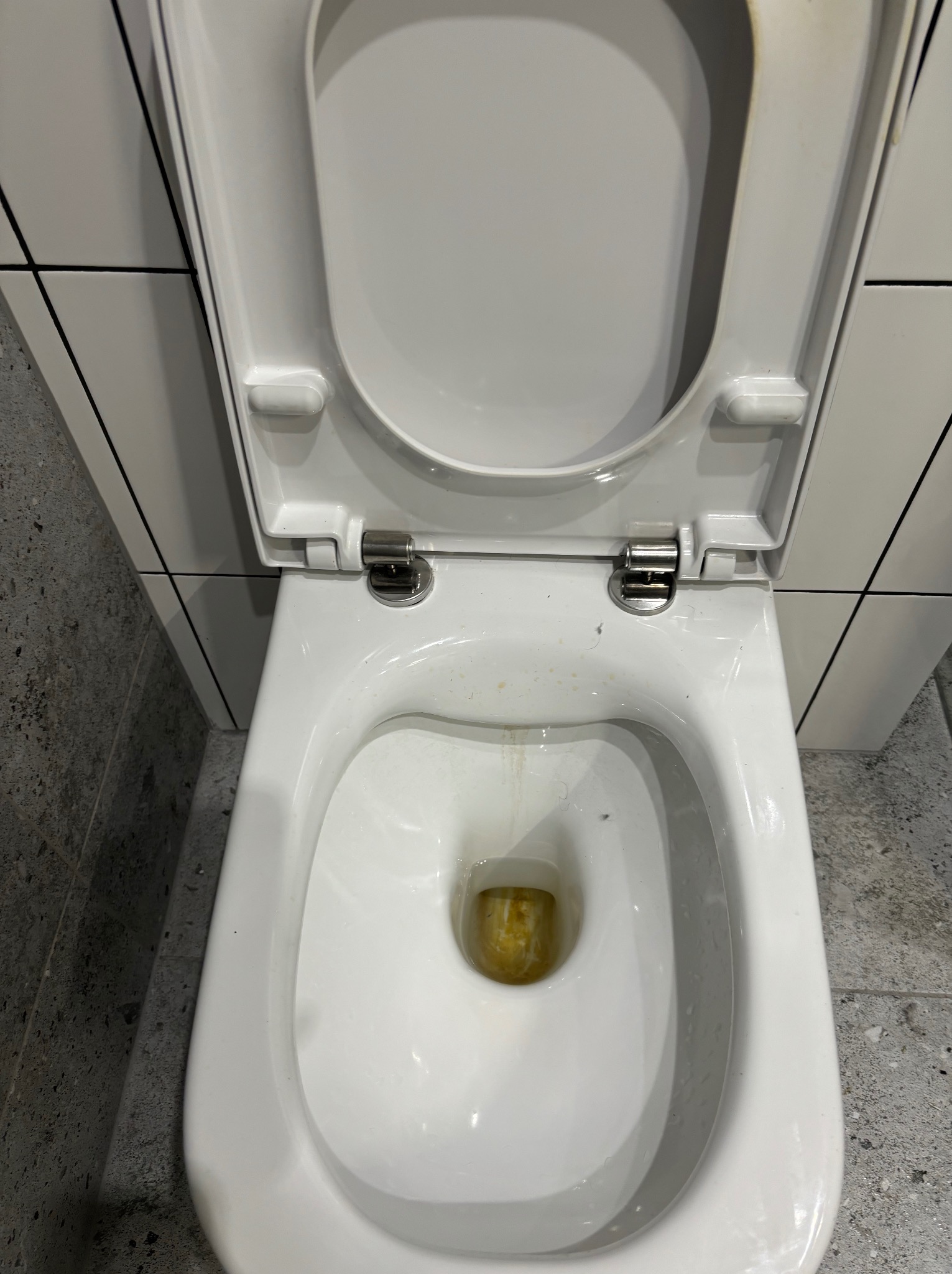 Otwarta, brudna, biała toaleta z widocznymi śladami użytkowania i osadami, na tle szaro-białych płytek.