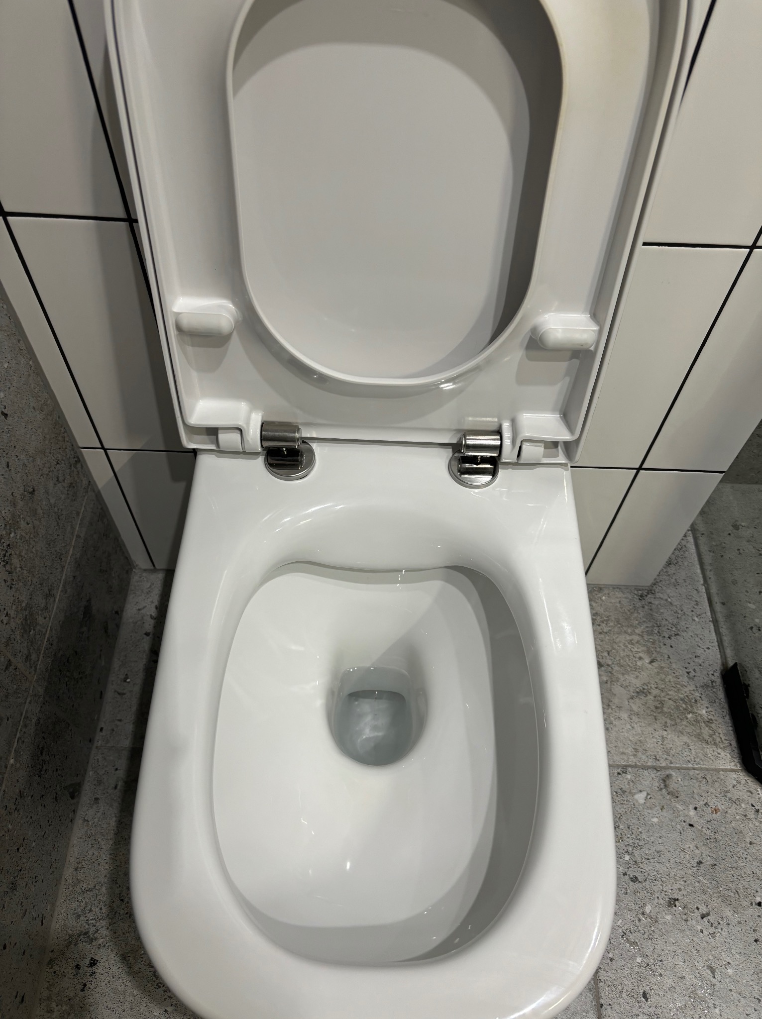 Widok z góry na otwartą, białą, wiszącą toaletę ze spłuczką, zamontowaną na tle białych płytek z czarnymi fugami i szarej posadzki.