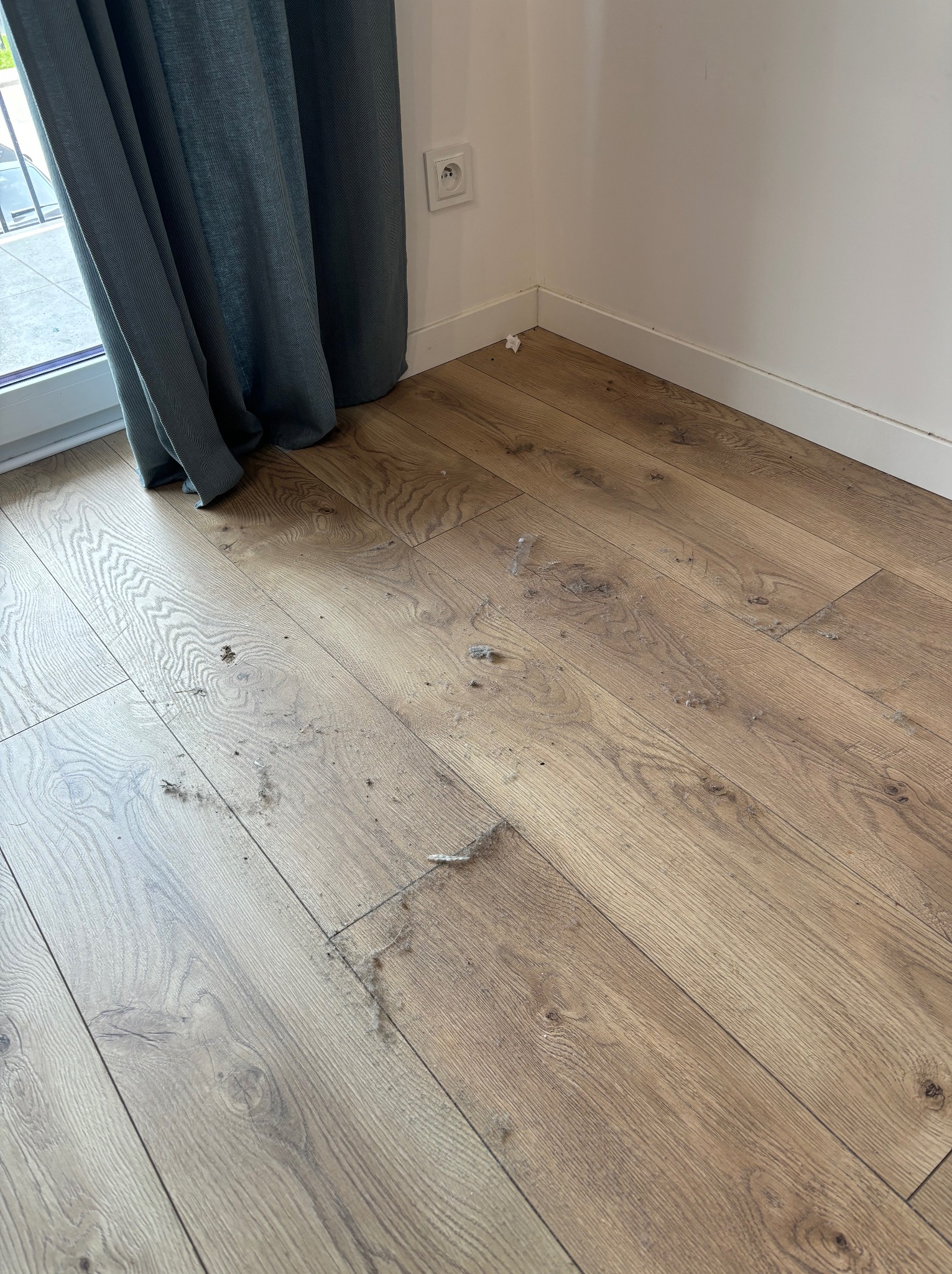 Drewniana podłoga z widocznymi kłębkami kurzu i okruchami, zasłonięta częściowo przez szarą zasłonę przy oknie, z gniazdkiem elektrycznym na białej ścianie w rogu pokoju.