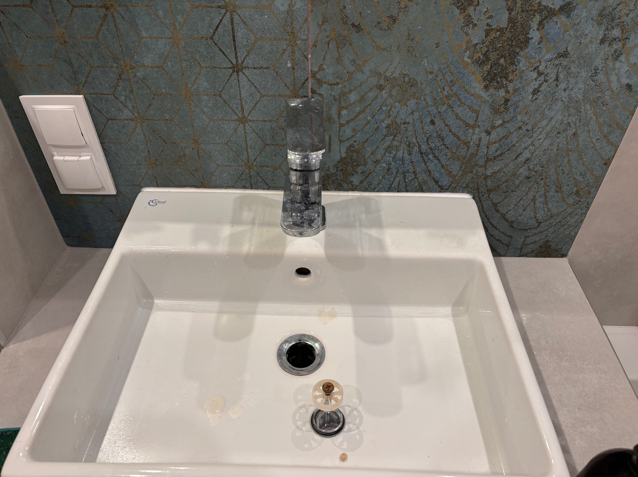 Biała umywalka z chromowaną baterią Ideal Standard i dekoracyjną armaturą odpływową na tle niebiesko-złotej ściany z geometrycznym wzorem i włącznikiem światła.