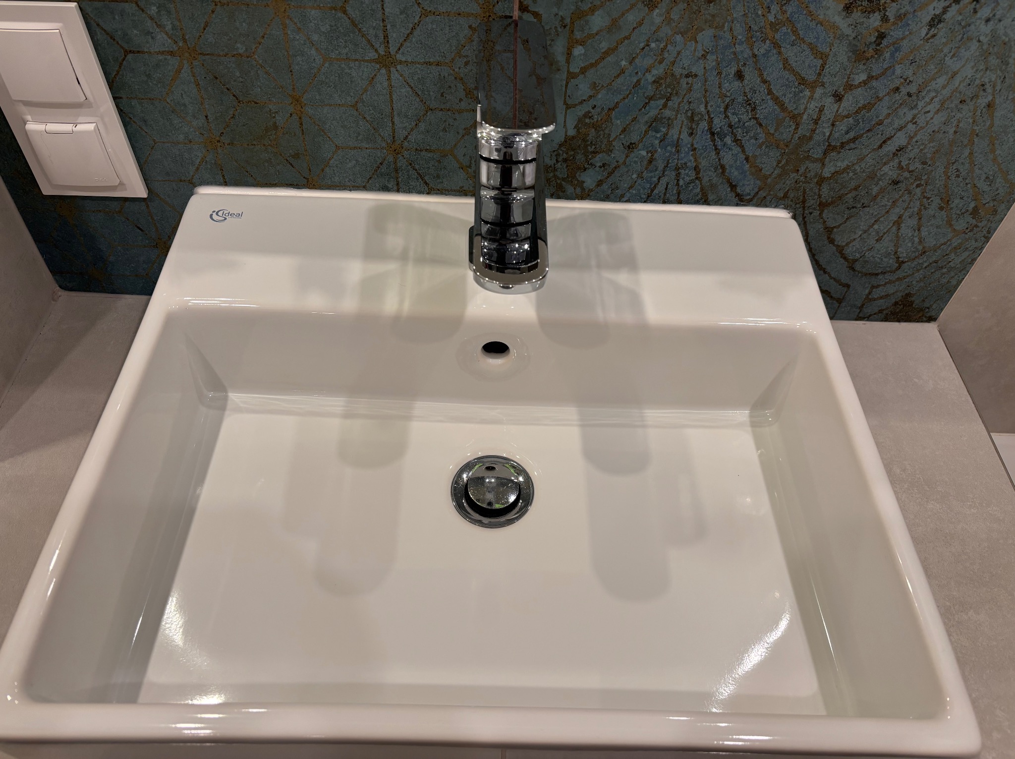 Biała, prostokątna umywalka z chromowaną baterią i odpływem, na tle wzorzystej, niebiesko-złotej ściany w łazience. Widoczny włącznik światła.