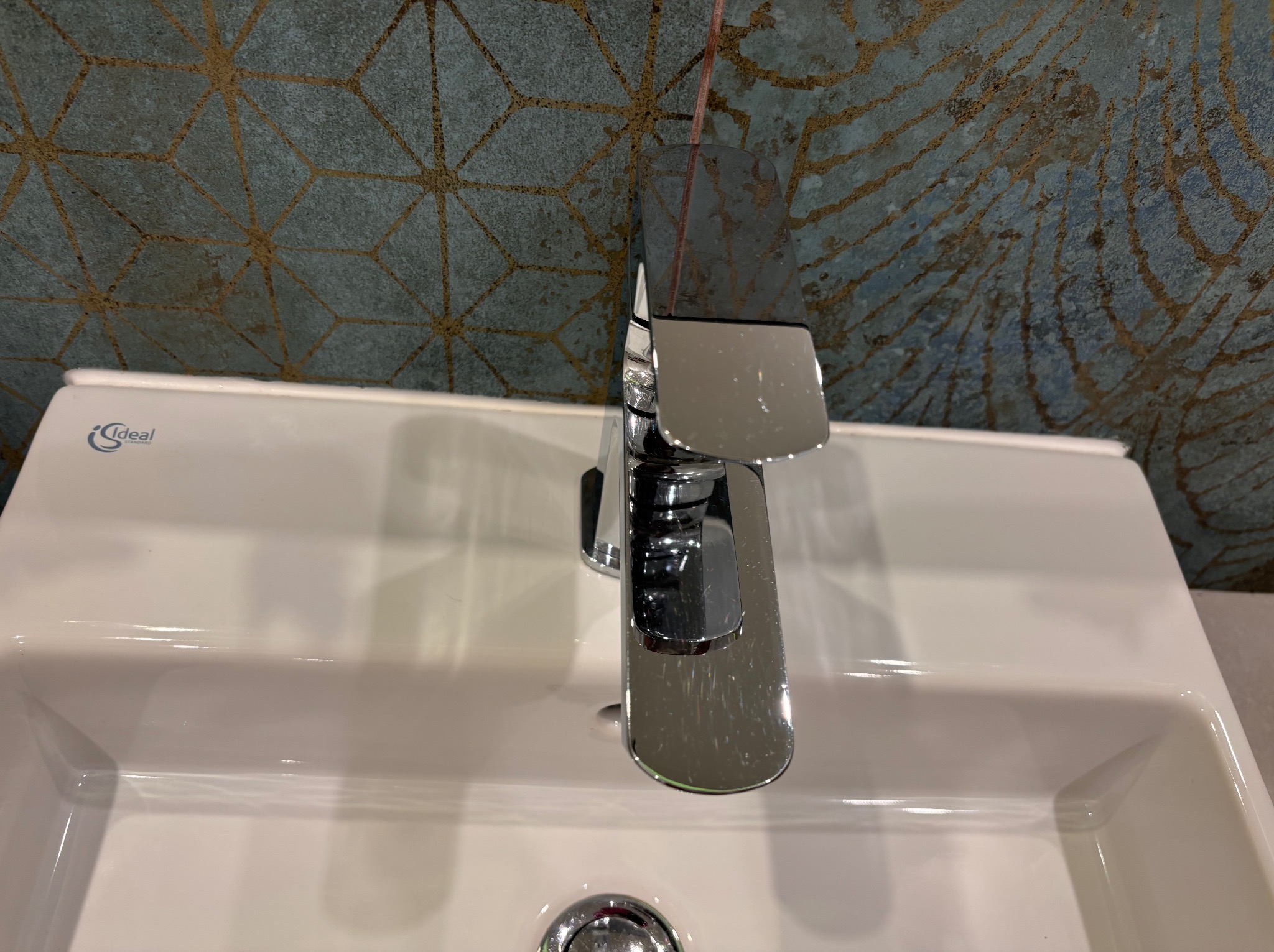 Bateria umywalkowa Ideal Standard na białej umywalce z dekoracyjną, niebiesko-złotą ścianą w tle.