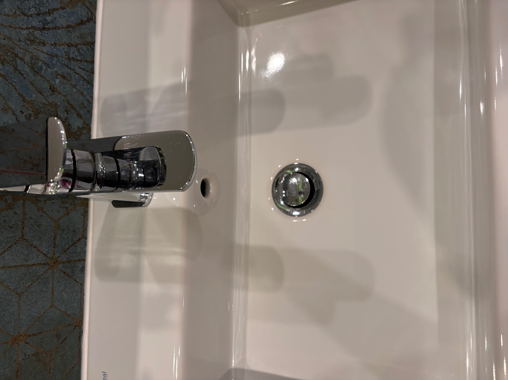 Biała umywalka z chromowaną baterią i korkiem spustowym na tle ciemnej, wzorzystej ściany.