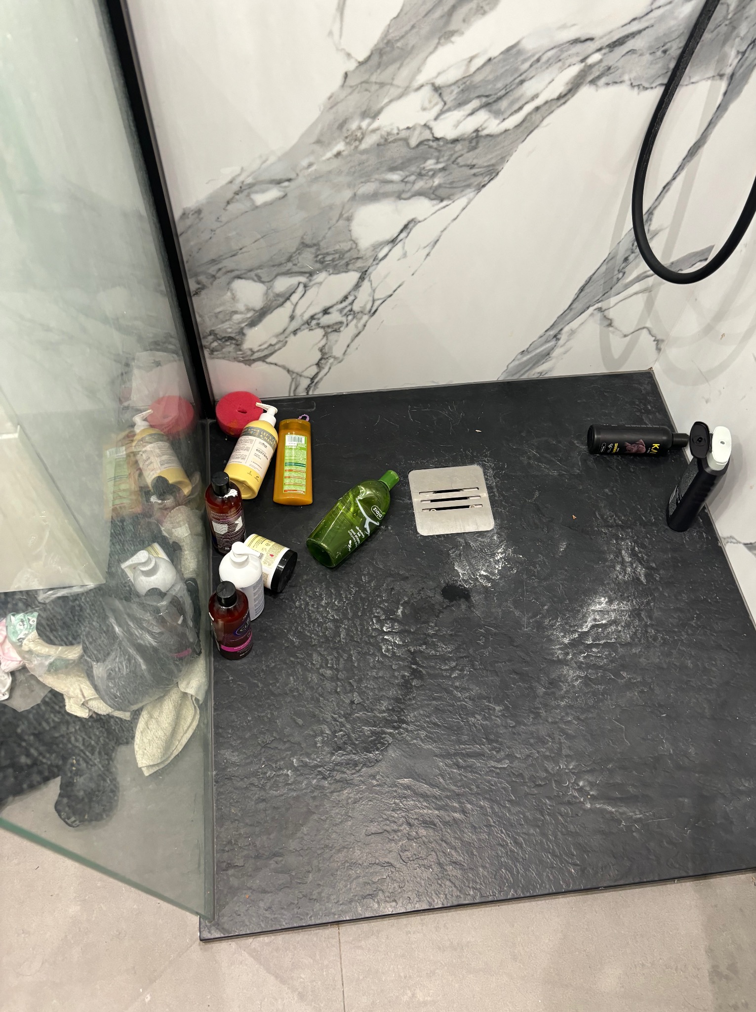 Czarny brodzik z odpływem liniowym w kabinie prysznicowej, otoczony kosmetykami i środkami czystości, marmurowa ściana w tle.