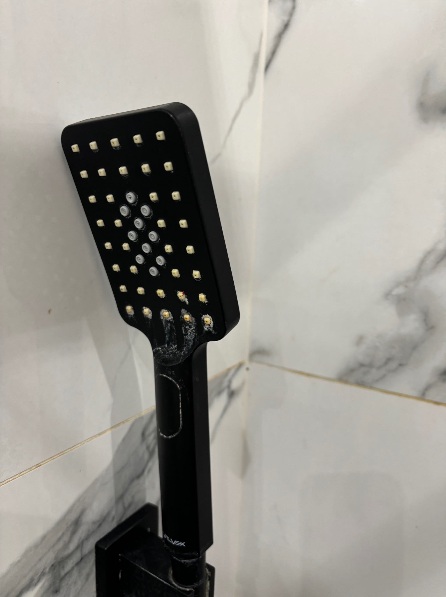 Czarna słuchawka prysznicowa z widocznymi osadami kamienia na tle marmurowej ściany.