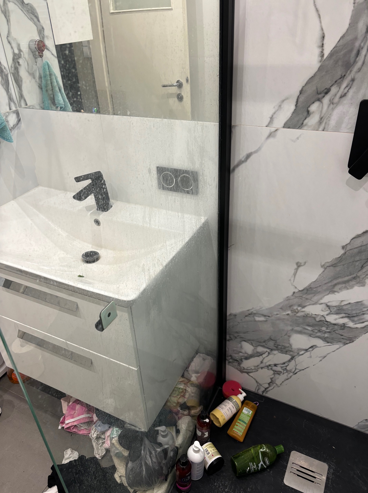 Zlew w łazience pokryty kroplami wody po umyciu, widoczne pozostałości po czyszczeniu, w tle marmurowe płytki i rozrzucone butelki środków czystości.