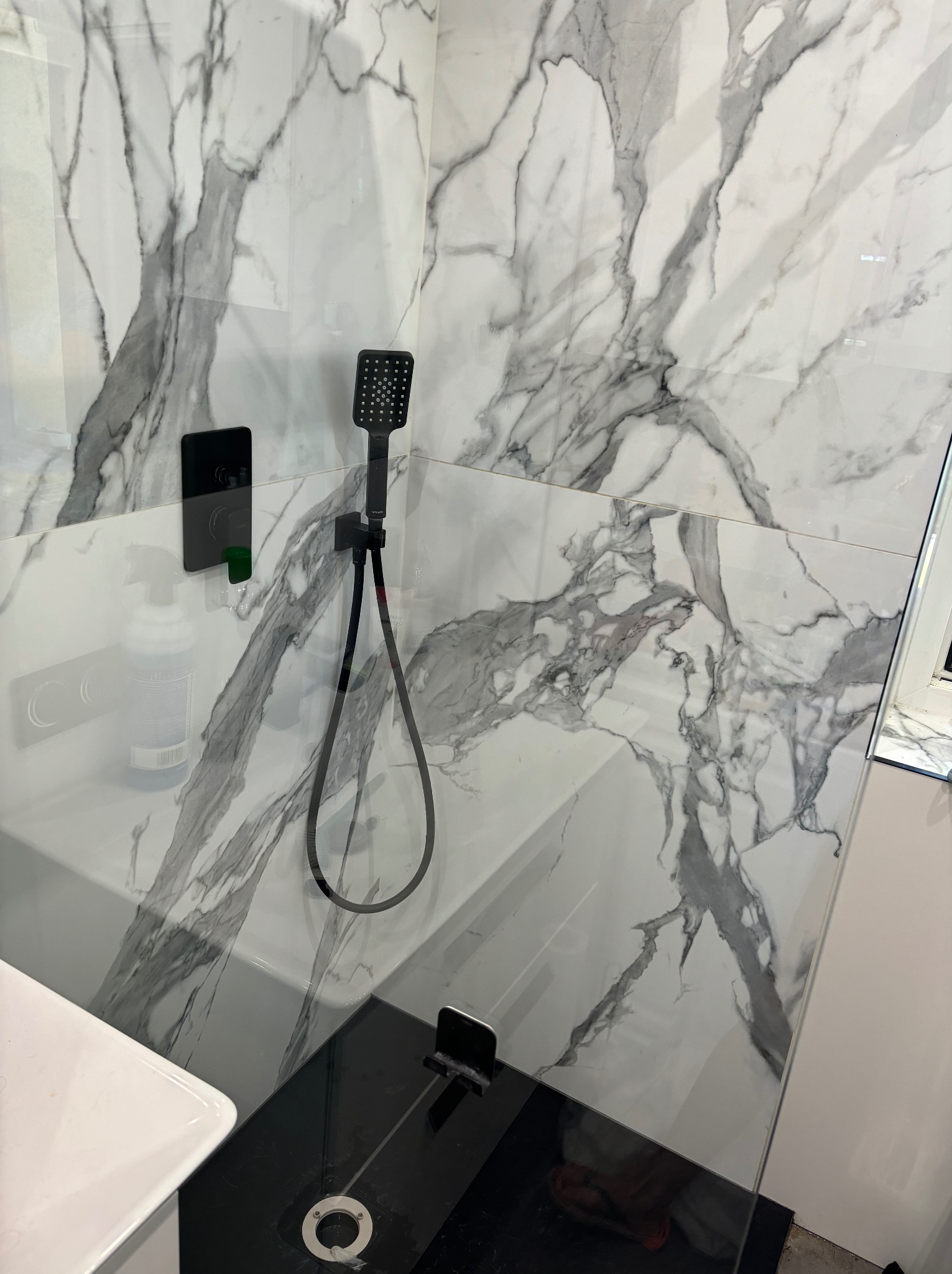 Nowoczesna kabina prysznicowa z czarną armaturą i białymi płytkami imitującymi marmur, widoczny dozownik mydła w płynie.