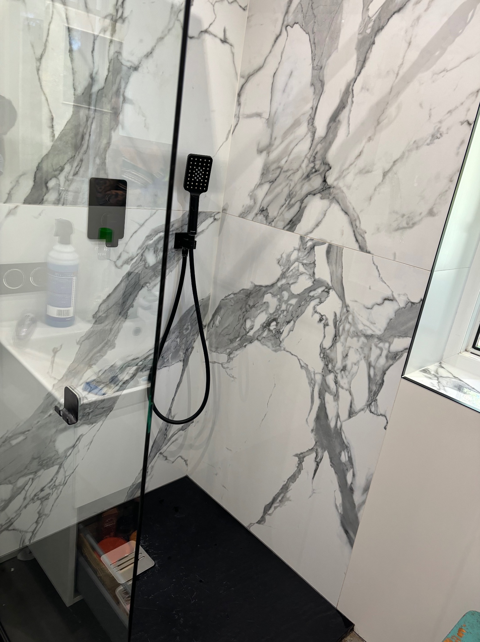 Nowoczesna kabina prysznicowa z czarną armaturą i marmurowymi płytkami w Krakowie. Widoczny czarny panel prysznicowy i półka z kosmetykami.
