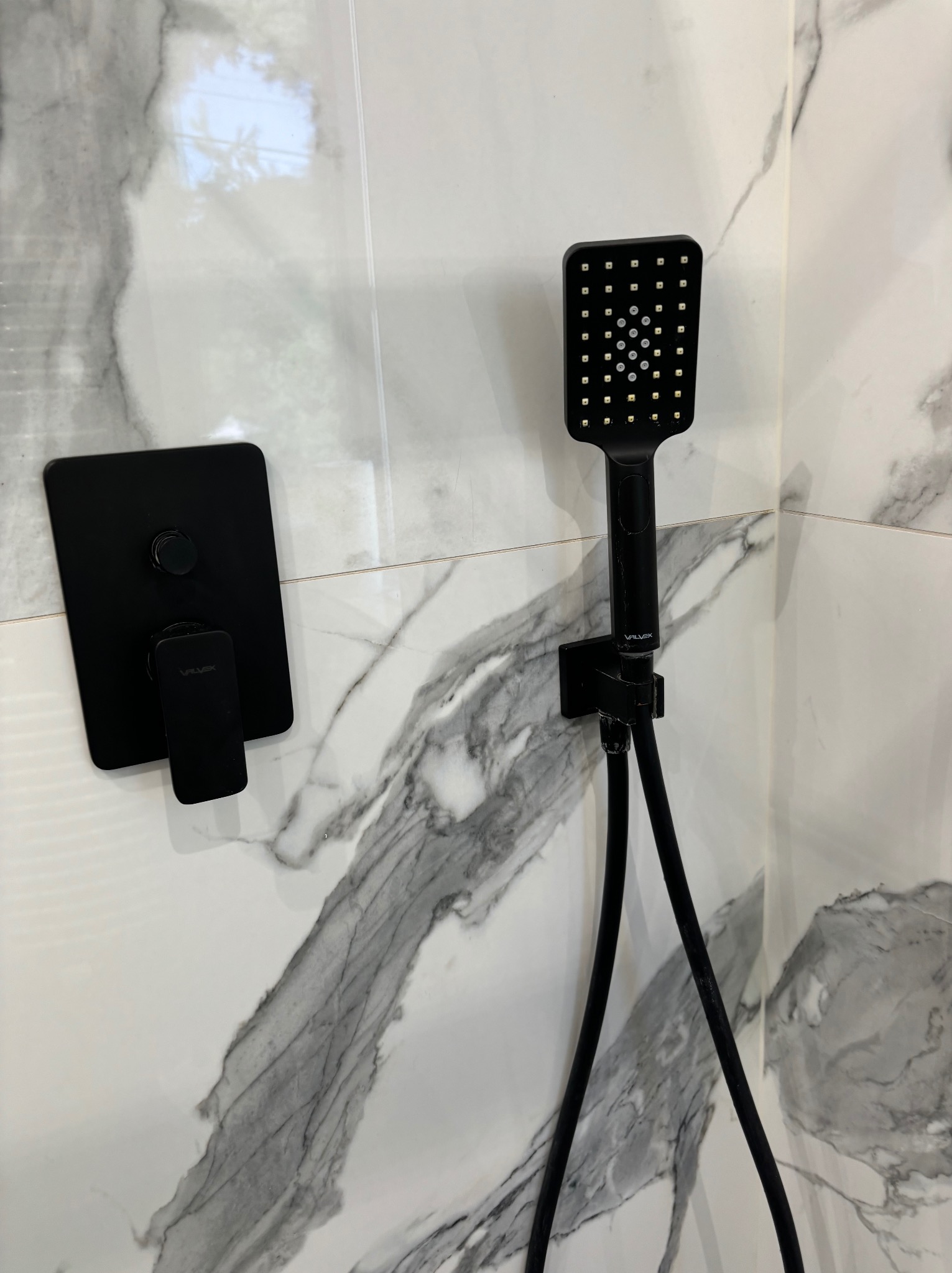 Czarny zestaw natryskowy z kwadratową deszczownicą i baterią podtynkową na tle białych płytek imitujących marmur.