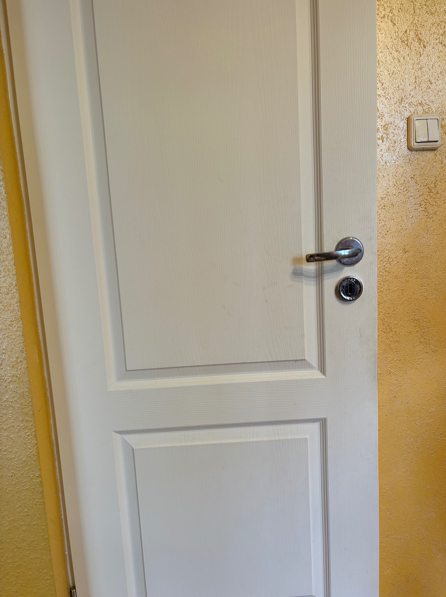 Białe, panelowe drzwi z chromowaną klamką i zamkiem, widoczne na tle ściany o fakturze pomalowanej na żółto, obok włącznik światła.