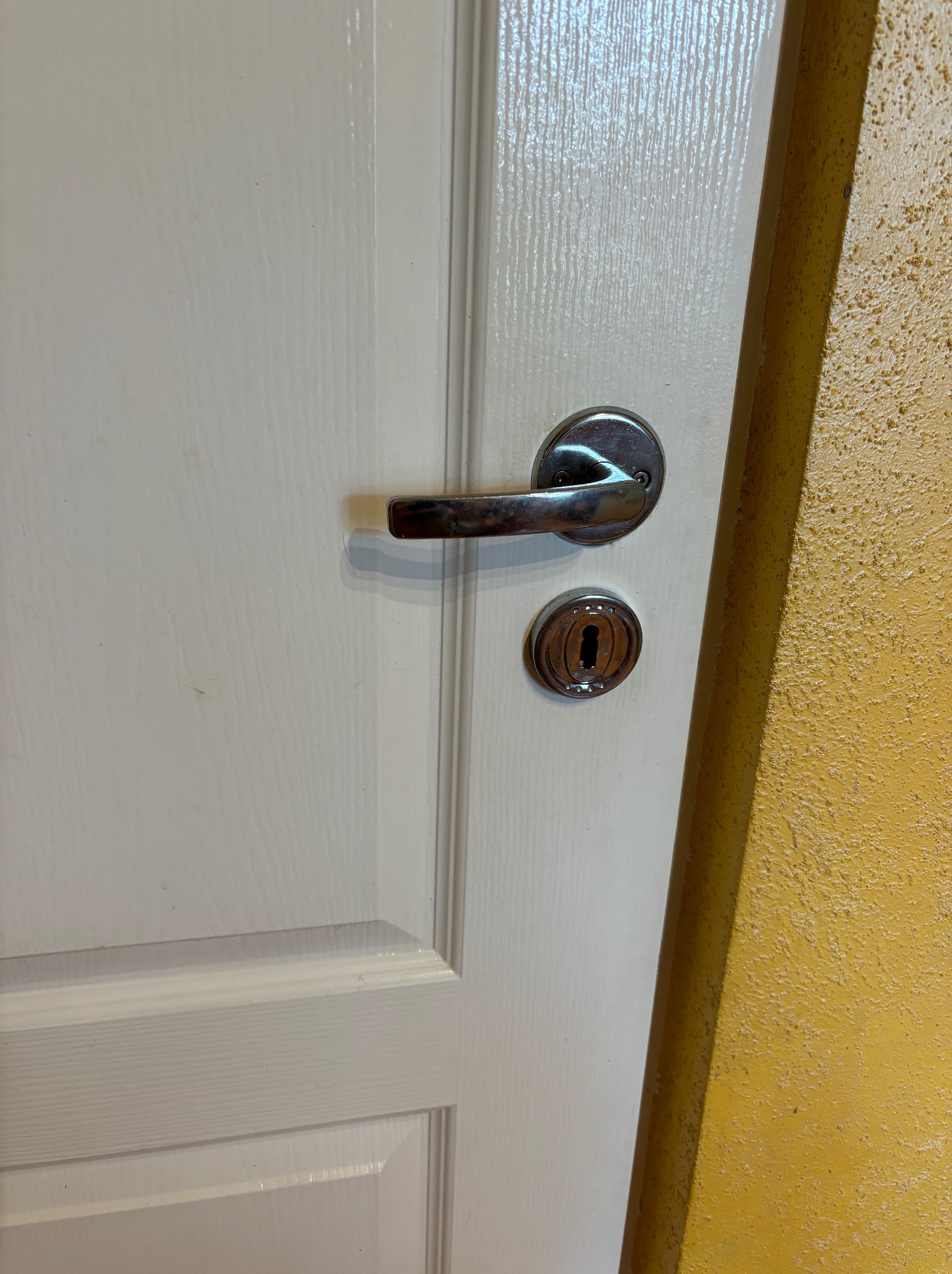 Białe drzwi z chromowaną klamką i zamkiem na tle żółtej, teksturowanej ściany.