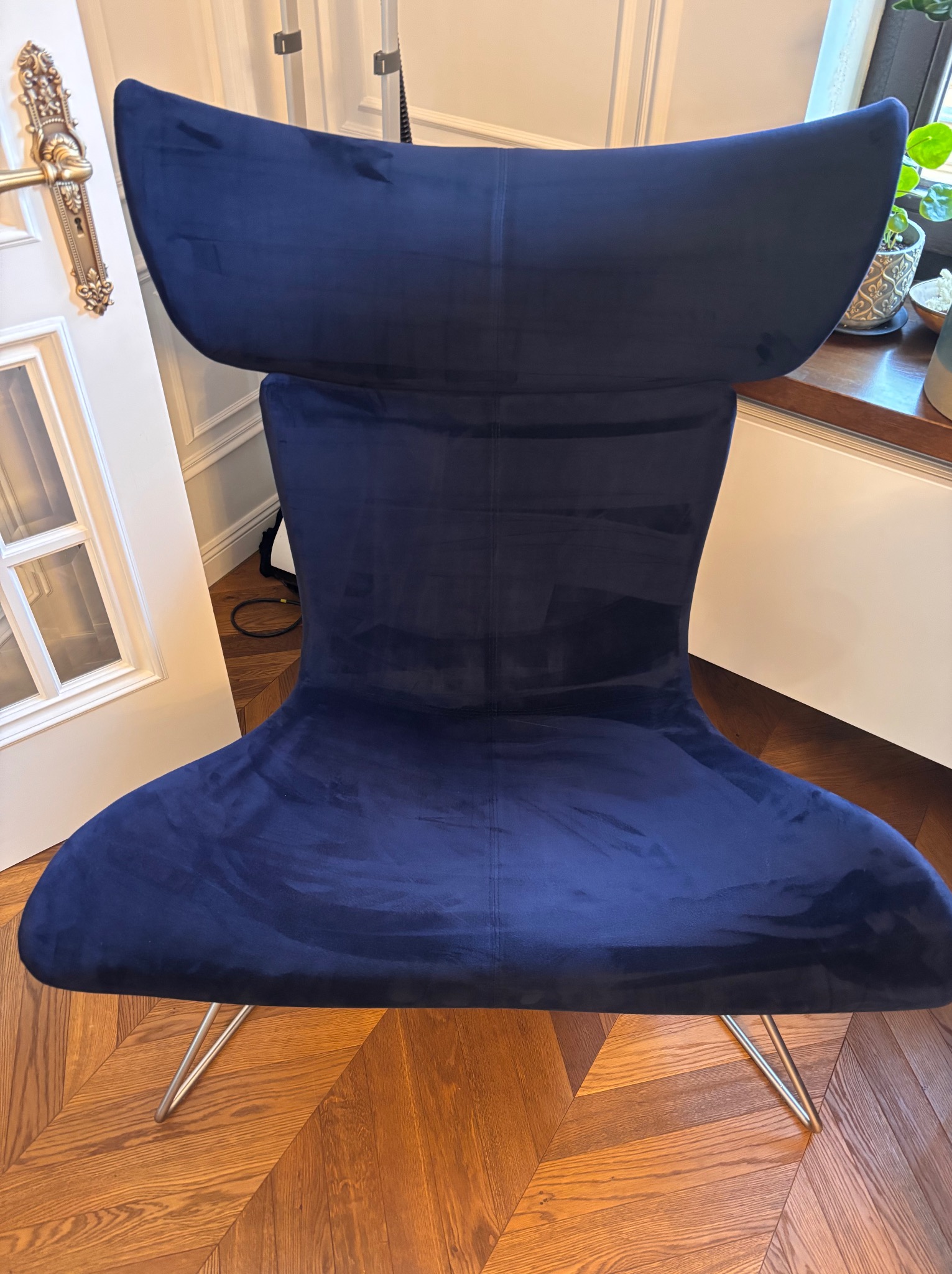 Eleganckie, granatowe krzesło tapicerowane welurem na metalowych nogach, ustawione na parkiecie w jodełkę, z fragmentem białych drzwi z ozdobną klamką i parapetem z roślinami w tle.