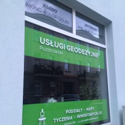 Okna biura geodezyjnego Puzdrowski z reklamą usług: podziały, mapy, tyczenia, inwentaryzacje. Widoczne odbicie budynku naprzeciwko.