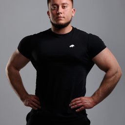 Pro Biznes Foto Rafał Wiśniewski - Mężczyzna w czarnej koszulce z logo marki, pozujący z rękami na biodrach na szarym tle.