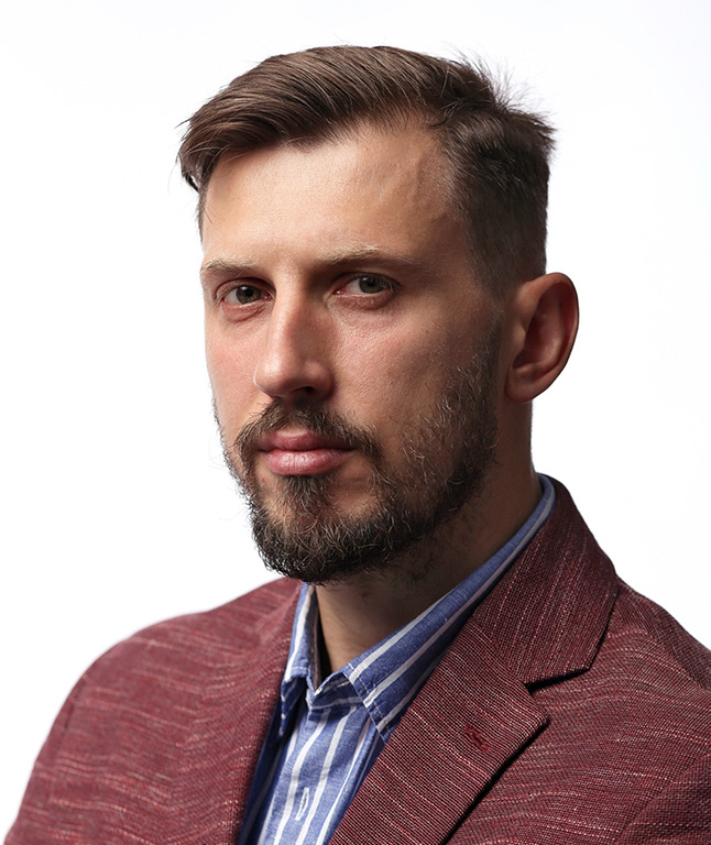 Portret mężczyzny z brodą w bordowej marynarce i koszuli w paski, na jasnym tle.