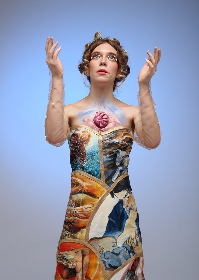 Portret kobiety w artystycznej sukni z motywami malarskimi i trójwymiarowym sercem na piersi, ręce uniesione w górę, włosy upięte z perłami, na jednolitym błękitnym tle.