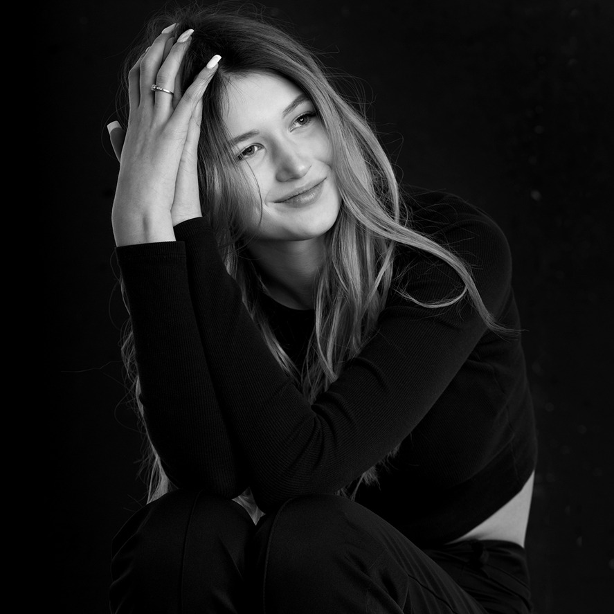 Portret młodej kobiety w czerni, z rękami uniesionymi do głowy, pierścionek na palcu, subtelny uśmiech, czarno-biała fotografia studyjna na ciemnym tle.