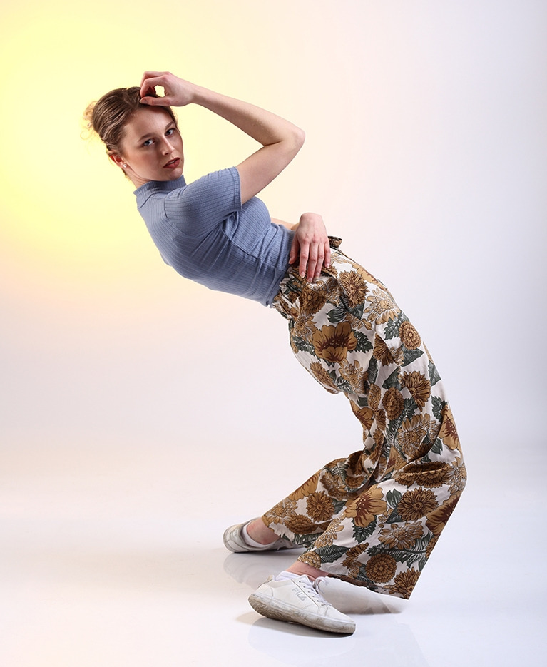 Młoda kobieta w niebieskiej prążkowanej bluzce i kwiecistych spodniach, pozująca w dynamicznej pozie na jasnym tle z żółtym gradientem, ubrana w białe, lekko zabrudzone sportowe buty.