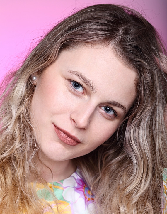 Portret młodej kobiety z blond włosami z delikatnym makijażem i perłą w uchu, na różowym tle.