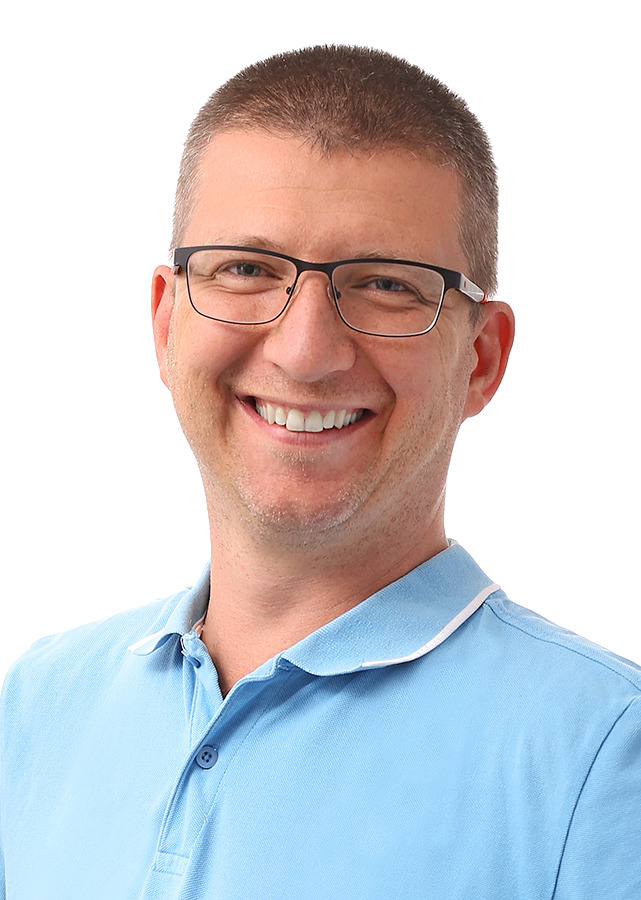 Portret uśmiechniętego mężczyzny w okularach z metalową oprawką i błękitnej koszulce polo z białym kołnierzykiem, na białym tle.