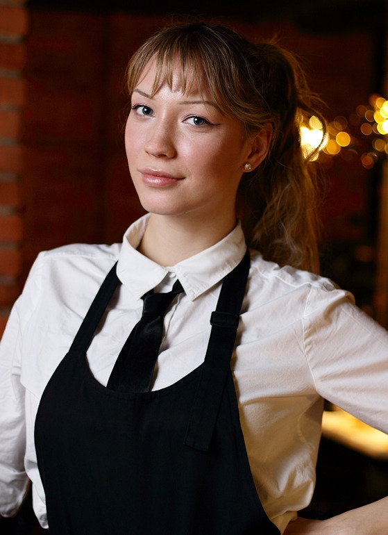 Portret młodej kobiety z blond włosami upiętymi w kucyk, ubrana w białą koszulę z czarnym krawatem i czarny fartuch, pozująca na tle ceglanej ściany i rozmytych świateł.