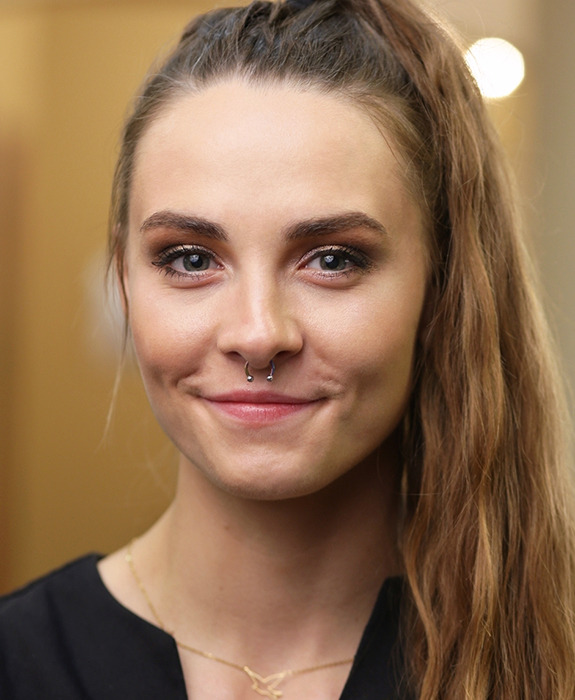 Portret uśmiechniętej kobiety z kolczykiem w przegrodzie nosowej, włosy spięte w wysoki kucyk, delikatny makijaż oczu i subtelna złota biżuteria na szyi.