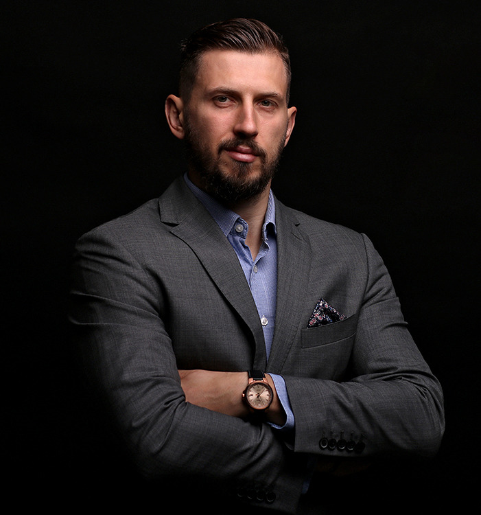 Portret mężczyzny w szarym garniturze, z brodą i eleganckim zegarkiem na tle ciemnego, niejednolitego tła.