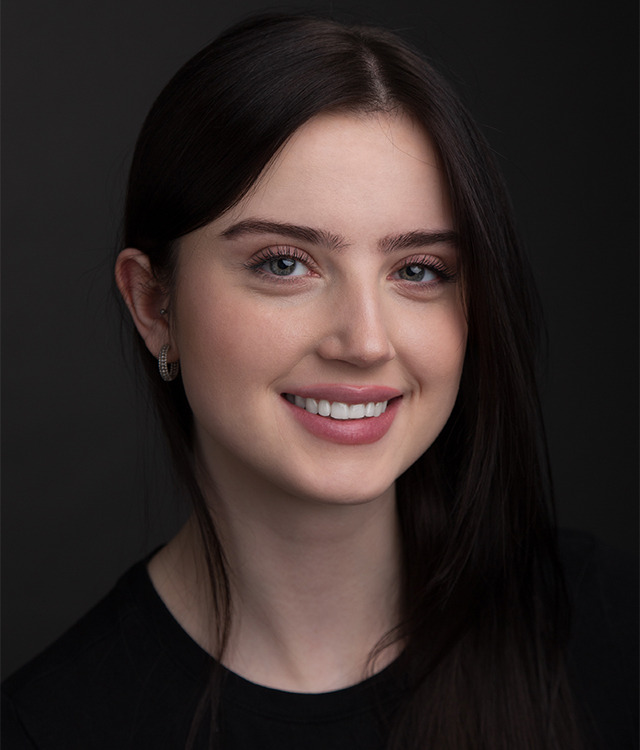 Portret młodej kobiety z ciemnymi włosami i jasnymi oczami, uśmiechniętej, z delikatnymi kolczykami w kształcie kół, na ciemnoszarym tle.