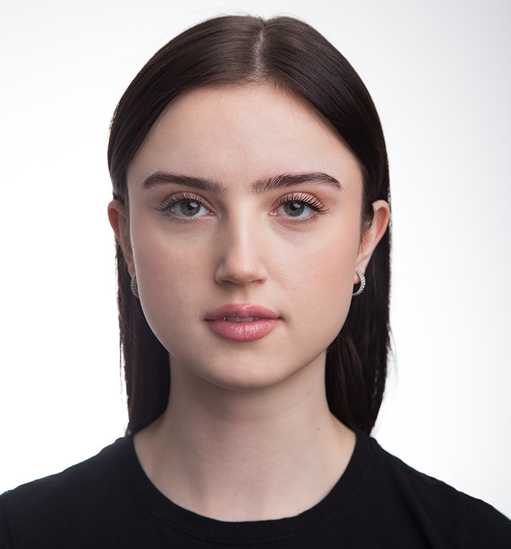 Portret młodej kobiety z ciemnymi włosami, skierowanej frontalnie, z delikatnym makijażem i srebrnymi kolczykami na białym tle.