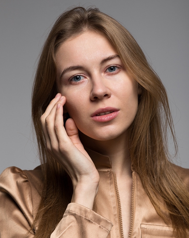 Portret kobiety z jasnymi włosami i niebieskimi oczami, w złotej bluzce, dotykającej policzka dłonią na szarym tle.