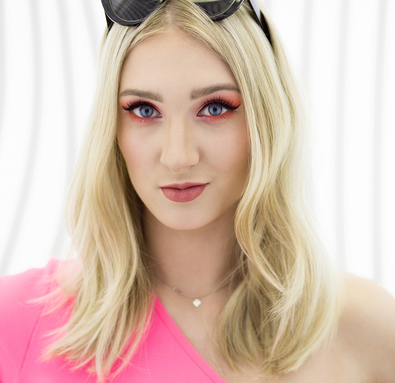 Portret kobiety z blond włosami, w różowej bluzce na jedno ramię, z intensywnym, pomarańczowym makijażem oczu i okularami na głowie, na jasnym tle.