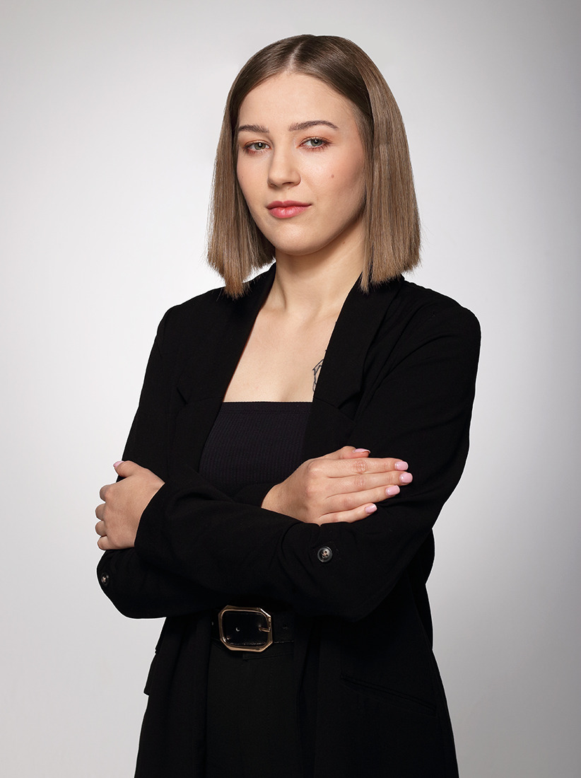 Portret kobiety w czarnym garniturze z paskiem i złotą klamrą, skrzyżowane ręce, neutralne tło, krótka fryzura, delikatny makijaż, studio.