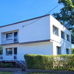 Rafał Lesik MAJSTER LESIK - Dwupiętrowy, biały dom z balkonami, otoczony zielenią i niskim, ozdobnym ogrodzeniem. Widoczne okna z brązowymi ramami i fragment instalacji na elewacji.