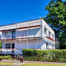 Rafał Lesik MAJSTER LESIK - Dwupiętrowy dom w trakcie malowania elewacji, otoczony rusztowaniem. Widoczne krzewy i drzewa w tle, błękitne niebo. Ujęcie w słoneczny dzień.