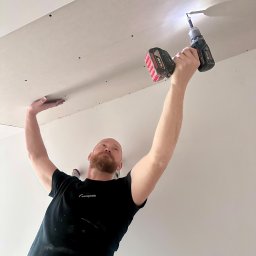 Rafał Lesik MAJSTER LESIK - Pracownik montuje płytę gipsową do sufitu za pomocą wkrętarki akumulatorowej. Widoczne otwory po wkrętach. Mężczyzna w czarnej koszulce z logo firmy.