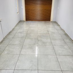 Rafał Lesik MAJSTER LESIK - Wykończone wnętrze garażu z szarą podłogą z płytek i brązową bramą rolowaną. Świeżo pomalowane ściany na szaro. Widoczne niedociągnięcia przy oknie.