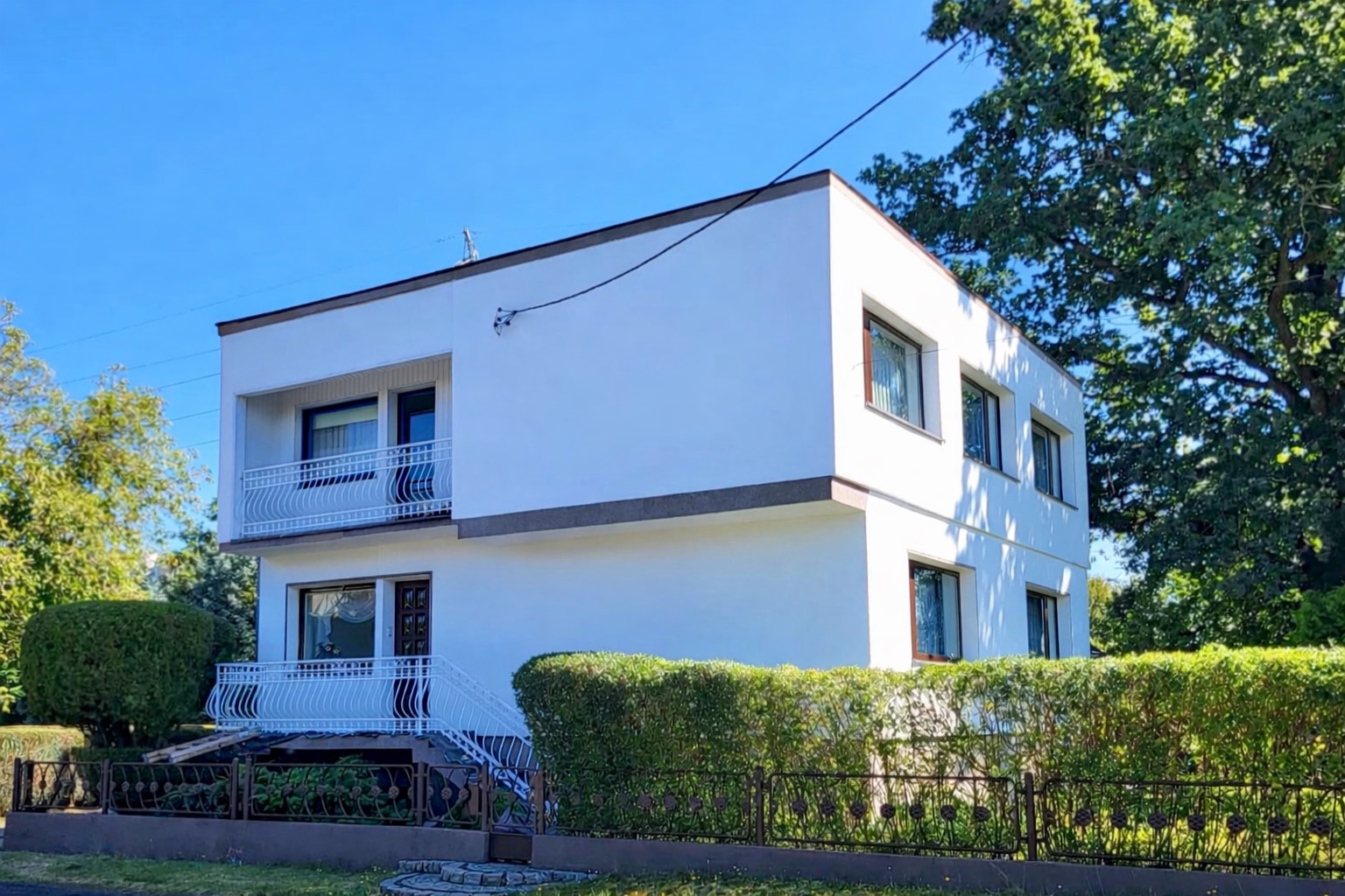 Dwupiętrowy, biały dom z balkonami, otoczony zielenią i niskim, ozdobnym ogrodzeniem. Widoczne okna z brązowymi ramami i fragment instalacji na elewacji.