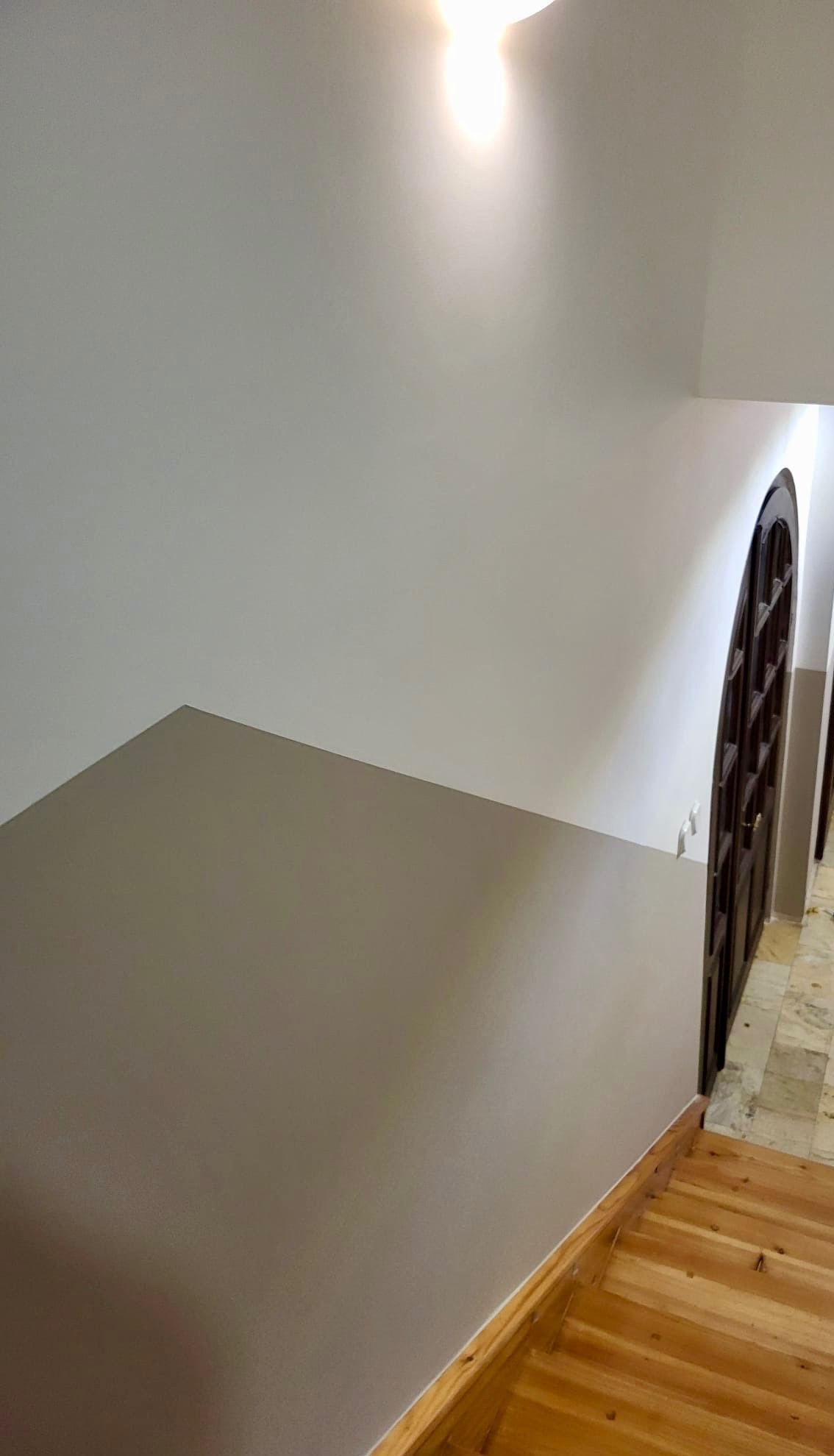 Ściana pomalowana na szaro, drewniane schody i ciemne, łukowe drzwi. Widok z góry, oświetlenie sufitowe. Minimalistyczne wnętrze po remoncie.
