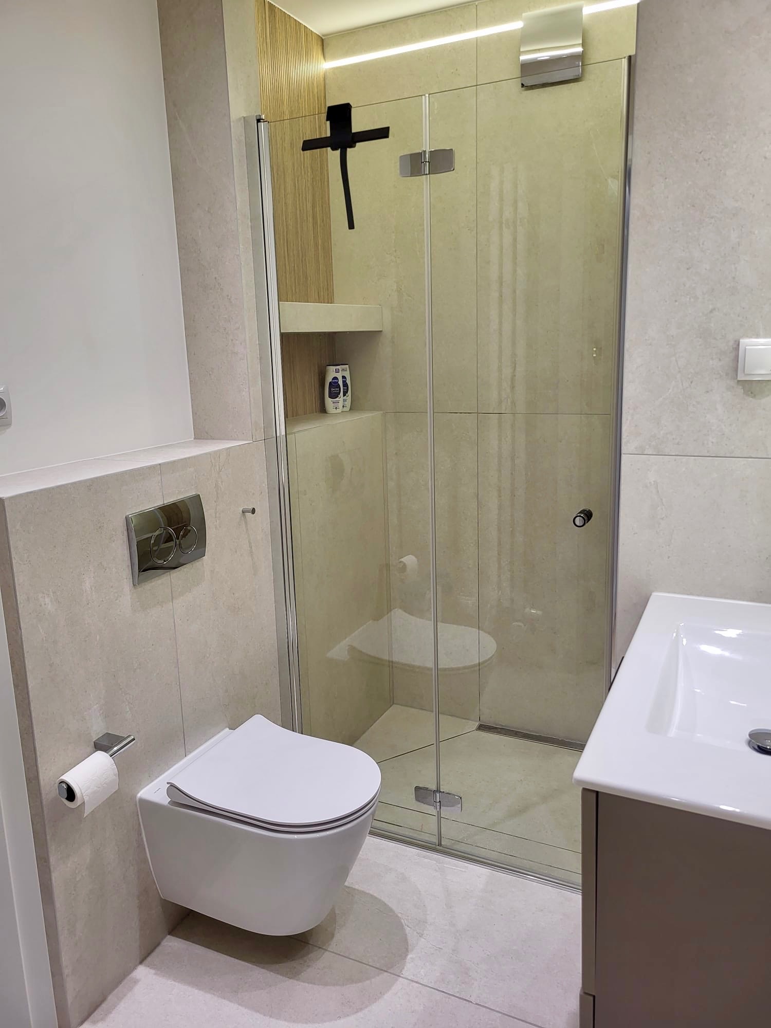 Nowoczesna łazienka z beżowymi płytkami, wiszącą toaletą i szklaną kabiną prysznicową. Minimalistyczny design z akcentami drewna i subtelnym oświetleniem LED.