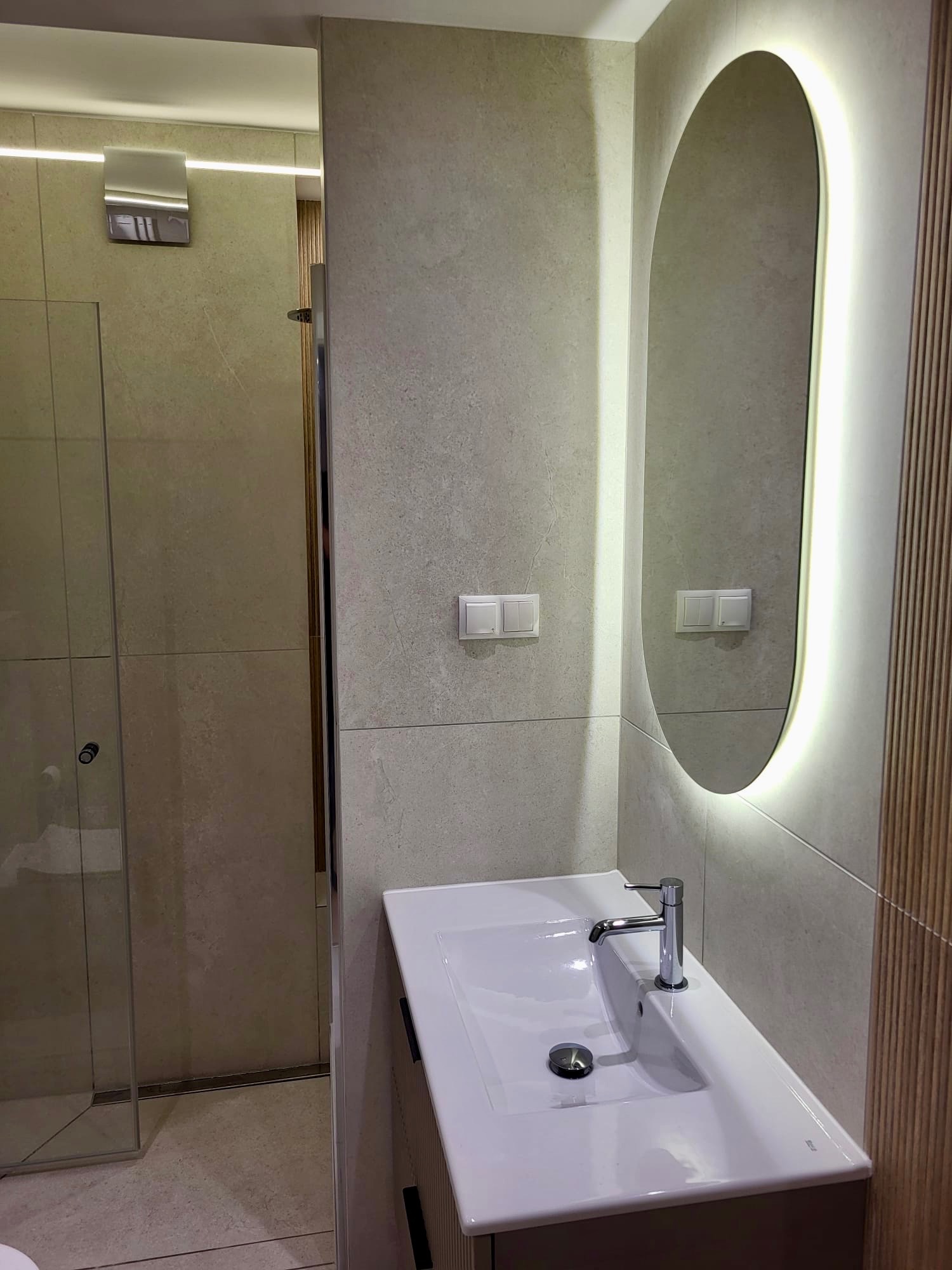 Nowoczesna łazienka z owalnym lustrem podświetlanym LED, białą umywalką nablatową i kabiną prysznicową. Płytki w odcieniach beżu i szarości. Minimalistyczny design.