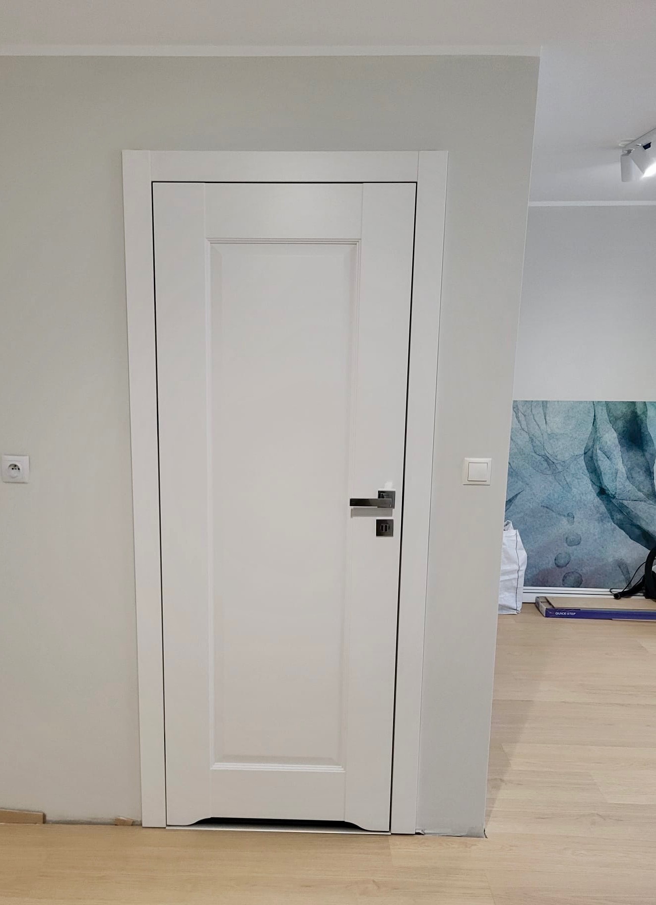 Białe, klasyczne drzwi wewnętrzne z srebrną klamką, zamontowane w jasnym pomieszczeniu z panelami podłogowymi i dekoracyjną tapetą w tle. Minimalistyczny design.