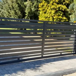 Nowoczesna, pozioma brama wjazdowa z metalowymi panelami w kolorze antracytowym, zamontowana na kostce brukowej, z widocznym stosem kostki brukowej obok, w tle zieleń drzew.