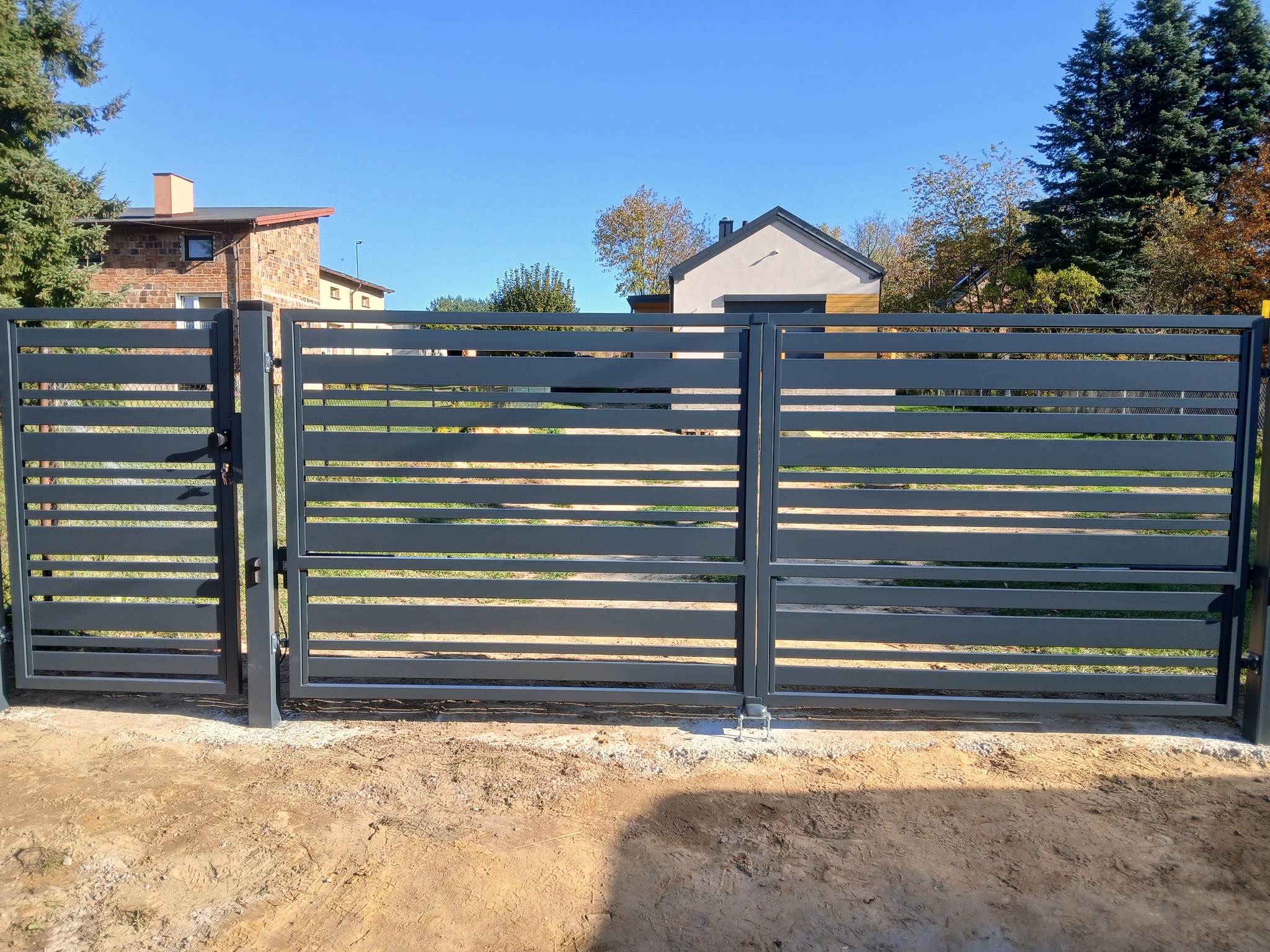 Nowoczesna, szara brama wjazdowa i furtka z poziomych profili stalowych, widoczne domy w tle, świeżo wyrównany teren.