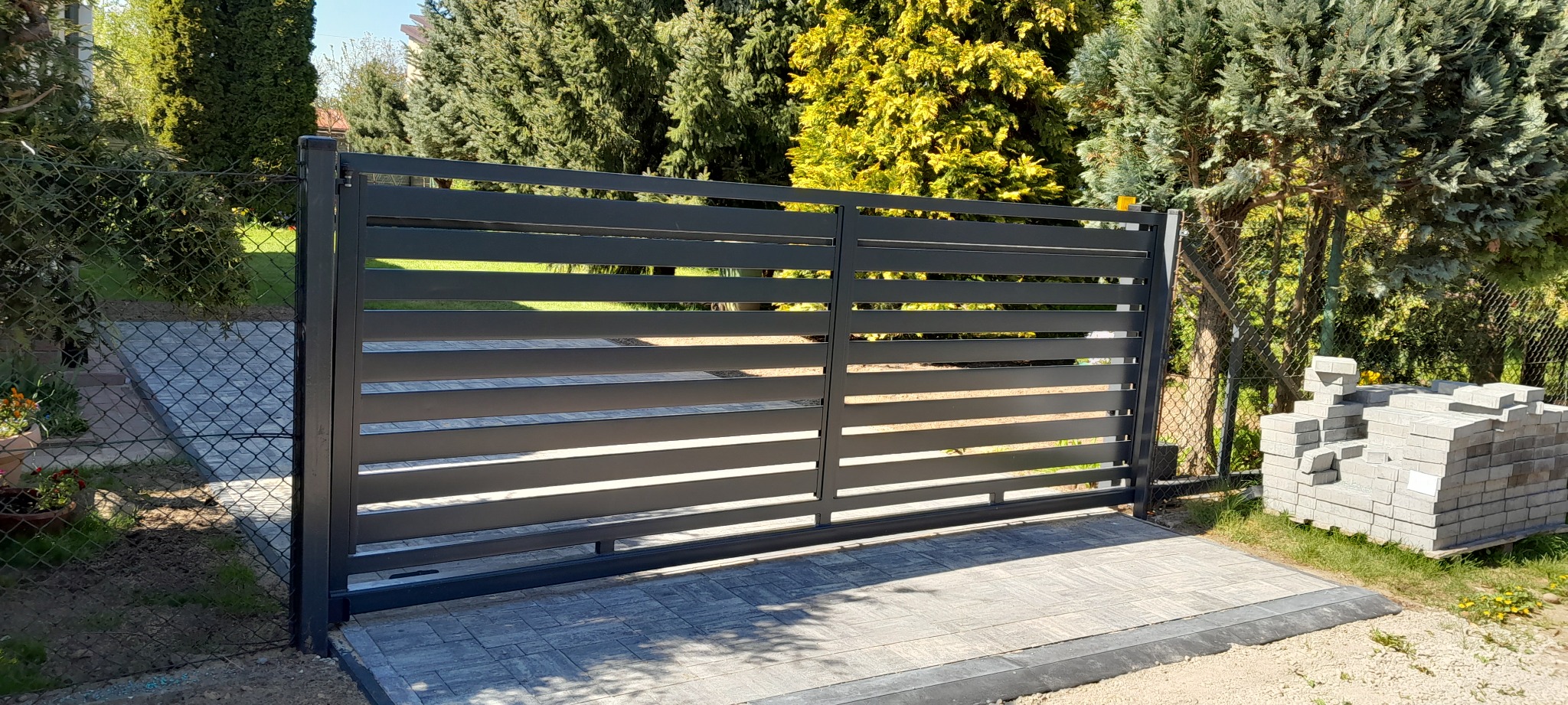 Nowoczesna, pozioma brama wjazdowa z metalowymi panelami w kolorze antracytowym, zamontowana na kostce brukowej, z widocznym stosem kostki brukowej obok, w tle zieleń drzew.