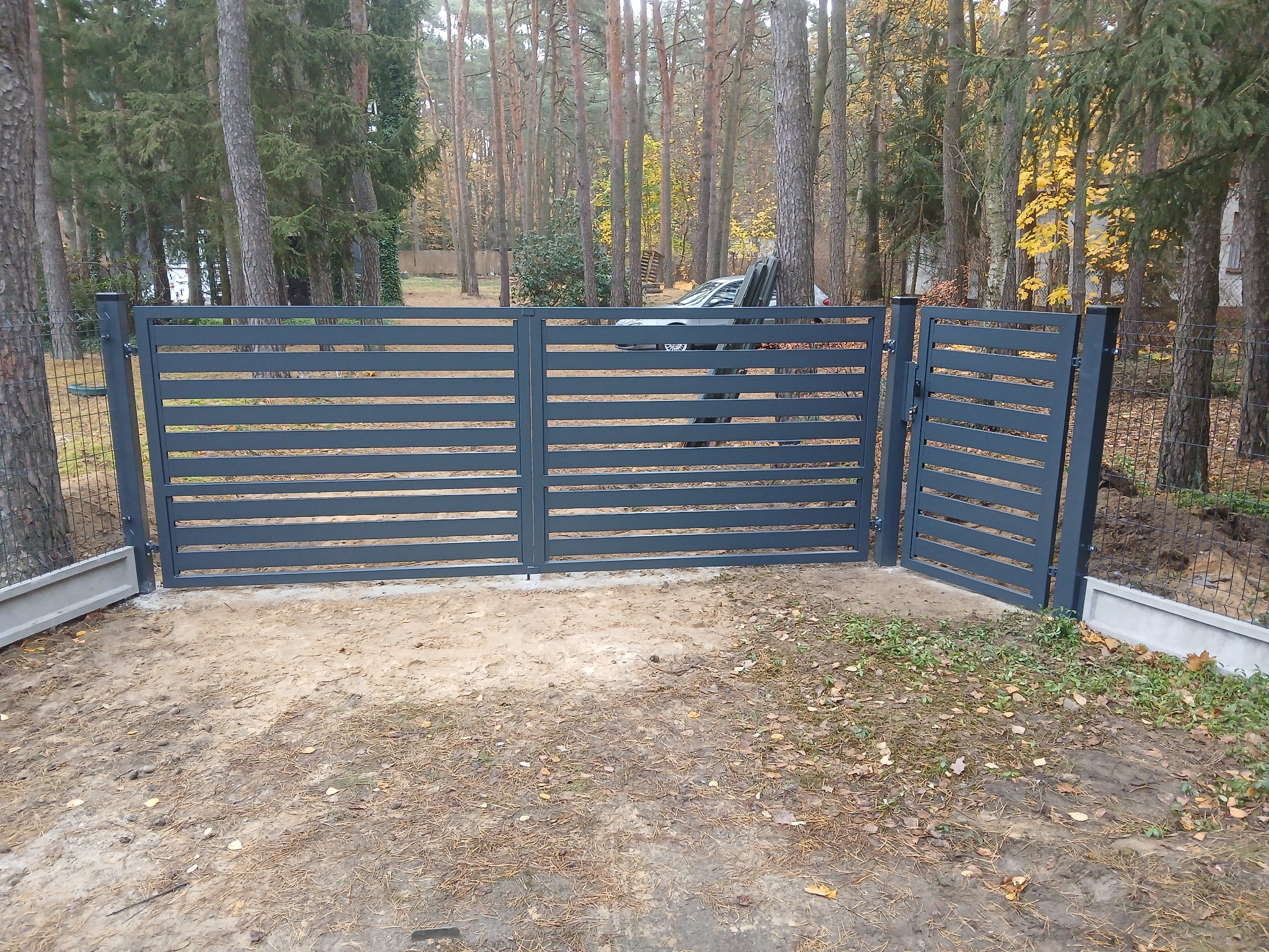 Nowoczesna, pozioma brama wjazdowa i furtka z metalowych paneli w kolorze grafitowym, zamontowana na posesji z widokiem na las sosnowy.
