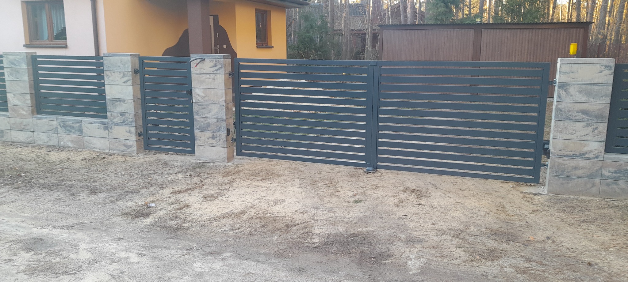 Nowoczesna, pozioma brama wjazdowa i przęsło ogrodzeniowe z szarego metalu, osadzone na słupkach z szarej kostki imitującej kamień, na tle domu z żółtą elewacją i lasu.